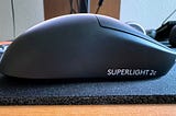 A close-up of the right side of a Logitech Superlight 2C gaming mouse, sitting atop a desk mat. The curve of the mouse’s smaller frame is visible, as well as its tweaked mouse wheel design. The words “Superlight 2c” are printed on its black plastic frame underneath the right-click button.