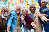 A joyful group of vibrant, stylish older adults smiling, laughing, and celebrating together against a bright blue background filled with colorful confetti and gold streamers.