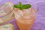 A cloudy, refreshing ginger-colored drink on ice, with a mint sprig and a lavender backdrop