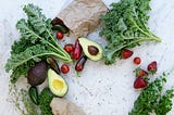 A white marbled countertop surface, with two brown bags placed on their side with fresh fruits and vegetables spilling out. There is a cluster of fresh green kale, sliced and whole avocados. Scattered around are stems of fresh herbs.