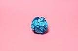A crumpled ball of blue paper against a solid pink background.