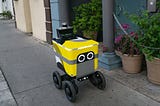 A photo of a Postmates food delivery robot going down a sidewalk.