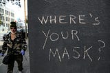 On the right side, three-quarters of the pic is a black exterior wall with “Where’s your mask?” written on it in chalk. The quarter of the photo on the left side is a person wearing latex gloves and a face mask, the latter under their chin.