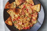 A wide dinner bowl of eggplant dip with toasted pita chips on it.