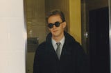 A sneering young man in a suit, black trenchcoat, and dark round sunglasses sneers into a mirror in a yellow-tiled restroom.