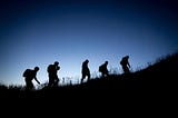 The silhouette of 5 people hiking up a hill, wearing backpacks.