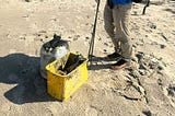 The author is carrying full gear and picking up trash on a beach with two containers in front of him.
