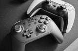A Nintendo Switch 2 Pro controller (left/bottom) and a white Sony DualSense controller sitting on a black desk mat.