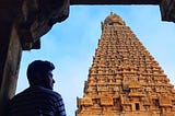 A man stands looking a temple structure in sand brown colour with blue sky behind.