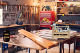 Workshop with organized tool wall and wooden workbenches in warm lighting. It has humorous signs including ‘DON’T USE THE YELLOW EXTENSION CORD IF IT’S RAINING ON A TUESDAY’, a ‘NASA SPECIAL’ bottle, and toolbox labeled ‘GOOD PLIERS’.