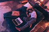 A young guy in checkered pajama trousers falling asleep with a slice of pizza in his hand in bed next to a pizza box and open laptop. He has the itis.