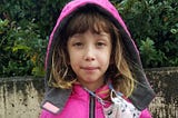 7-year-old kid in a pink hoodie with a mask around her neck holds her new Covid-19 vaccine card and wears a vaccinated sticker