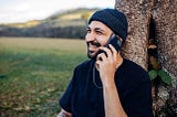 A bearded man in a black shirt and beanie sits in a field with his back to a tree, smiling and talking on a cell phone