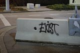 A piece of graffiti on concrete. It reads: “Exist”