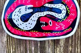 Color vertical image of a brightly colored glossy sticker laying at the bottom half od the frame, upon on a a wooden plank with very rounded like a dark wood ‘moon’ knot above it. The horizontal sticker of two yin-yang-ish wound snakes where a larger dayglo pink snake is wrapped fully around a smaller white snake in the middle of the sticker, all black background and outlined in white.