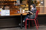 Older woman wearing face mask reading a book outside a cafe.