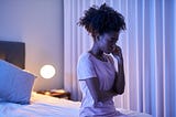 A troubled black woman sits at the edge of her bed.