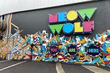 The side of a building featuring the words “Meow Wolf” in bright colors, and “You Are Here” below that, surrounded by a painted mural