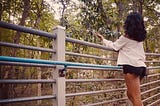 A woman admiring a leaf attached to a tree branch hanging over a pedestrian bridge in the middle of a community park