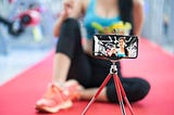 A photo of a woman recording herself doing a workout using her smartphone on a tripod.