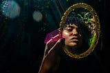 Young Black woman holding up and looking into a gilded mirror in the forest.