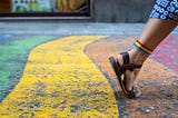 A woman’s foot with sandals and a rainbow flag tied to the knuckle making a step forward on a rainbow sidewalk.
