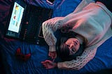 A woman is lying on bed with a gaming laptop and console.