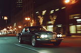 “Classic black luxury car driving through a bustling city street at night, its headlights illuminating the road. Motion blur emphasizes speed and sophistication, while the urban skyline with tall buildings and streetlights enhances the scene’s dynamic energy. Perfect for showcasing timeless elegance and high-end automotive design in a modern city setting.