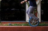 A man rides a blue unicycle atop a low brick wall