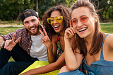Friends apparently sitting in a park smiling into a camera. Two of them are holding up two fingers. On the left a bearded man with a reversed baseball cap. In the middle and on the right two young women wearing coloured sunglasses.