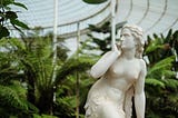 A marble statue of a thin woman looking off to the side, angrily and mistrustfully. The statue stands in front of lush ferns.