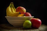 A still-life photograph of a bowl of fruit, with bananas, red apples, and a lime