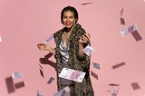 Woman with short hair and red lipstick wearing a silky lilac dress and a leopard print overcoat. She’s standing in front of a light pink background. Money is raining around her. 100 euro nots to be exact. She’s smiling and standing in a celebratory pose.