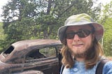 The author is wearing a tie-die shirt and hat. His beard and mustache are beginning to turn white. He’s wearing sunglasses and is standing in front of a rusty abandoned vintage car in the middle of the woods.