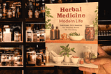 Background: Dark shelf with jars full of herbs. Foreground: Book cover of Herbal Medicine for Modern Life by Ruth A. Blanding, held by Gem Blackthorn