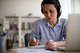 A woman wearing headphones and working on her laptop takes notes.