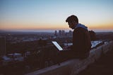 man on a ledge with his laptop at the rooftop overlooking the city sunrise