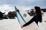 three australian accent variations girl surfer holding surfboard on beach looking out at water sand and rocks smiling