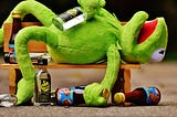 Kermit The Frog lying on wooden bench surrounded by glass bottles. Kermit appears drunk with a bottle of clear bottle presumed to be vodka in one hand and a brown bottle presumed to be beer in the other. He’s lying on his back with his mouth open and one leg hanging over the back of the bench.