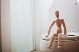 A wooden artist’s model is posed sitting on a toilet