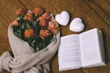 Roses wrapped in a cardigan, a book, and two white ceramic hearts rest on a table.