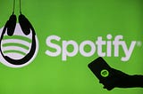 Headphones hang upside down as a hand holds a phone with the Spotify logo displayed in front of a screen showing Spotify