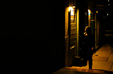 A person outside of a bar at night, illuminated by warm light, the other half of the frame cast in shadows.