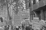 A black-and-white photo captures a pro-immigration rally with a crowd of people holding various signs. The signs display messages like “Refugees Welcome,” “No one is born hating another person,” and “Teach British colonial history.” The background features a historic building with detailed architecture, as well as trees and lampposts. The mood is one of peaceful activism, with people gathered to promote tolerance and acceptance of refugees. (AI-generated description.)