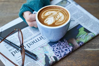 A hand holding a cup of coffee over a newspaper with a pair of glasses and a pen.