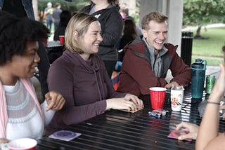 Three people site at a metal picnic table. There are cards on the table and they are smiling about to play a game.