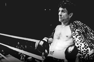 A black and white still of Robert De Niro leaning against a boxing ring. He’s wearing boxing gloves and shorts and has a leopard print cape over his shoulders.