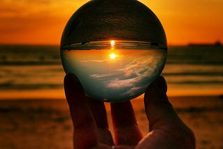 Someone holds a sphere in their fingertips against a sunset over the ocean.  The sphere reverses the image making the sky look like it's in the ocean and the sand from the beach look like it's in the sky. 