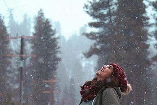 A dark-haired woman smiles at the sky as snowflakes fall around her. She is surrounded by trees
