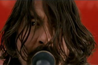 Close-up of Dave Grohl’s face singing into a microphone, hair covering his face, from the music video for “The Pretender.” Dave has a wall of pure red behind him.